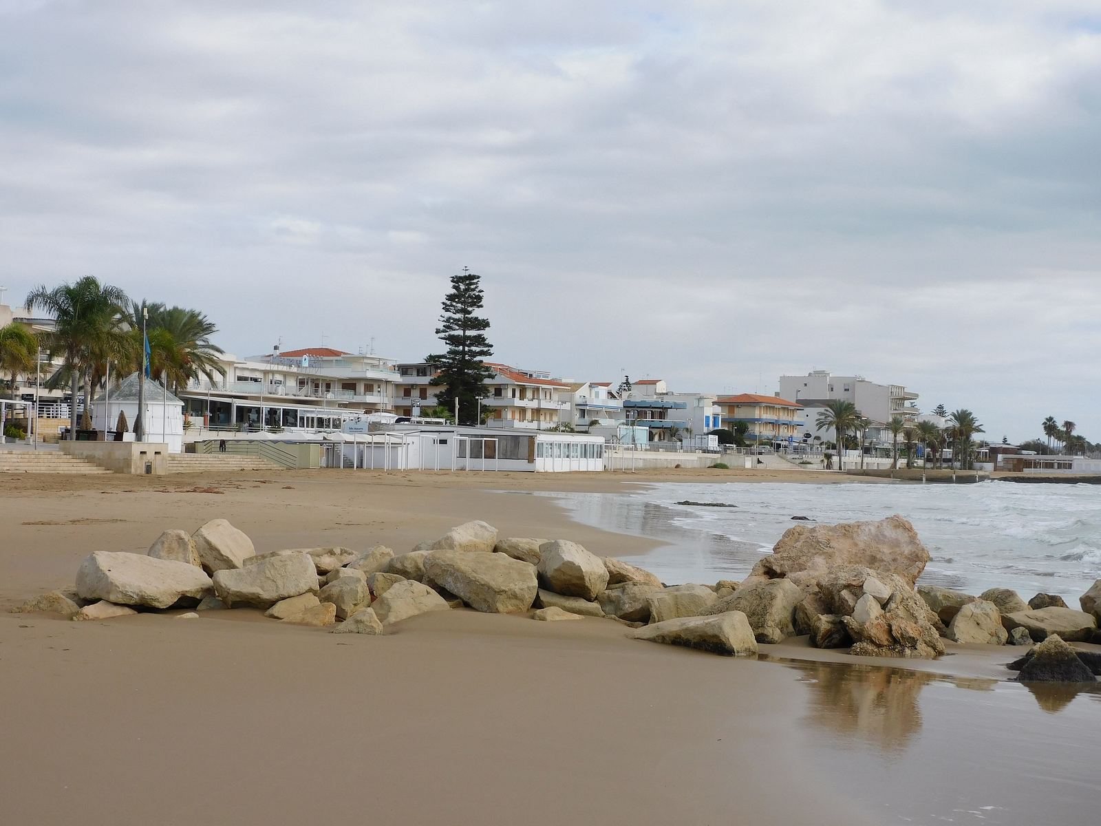 Spiaggia di Marina di Ragusa con scogli sulla battigia, palme e case sul lungomare.