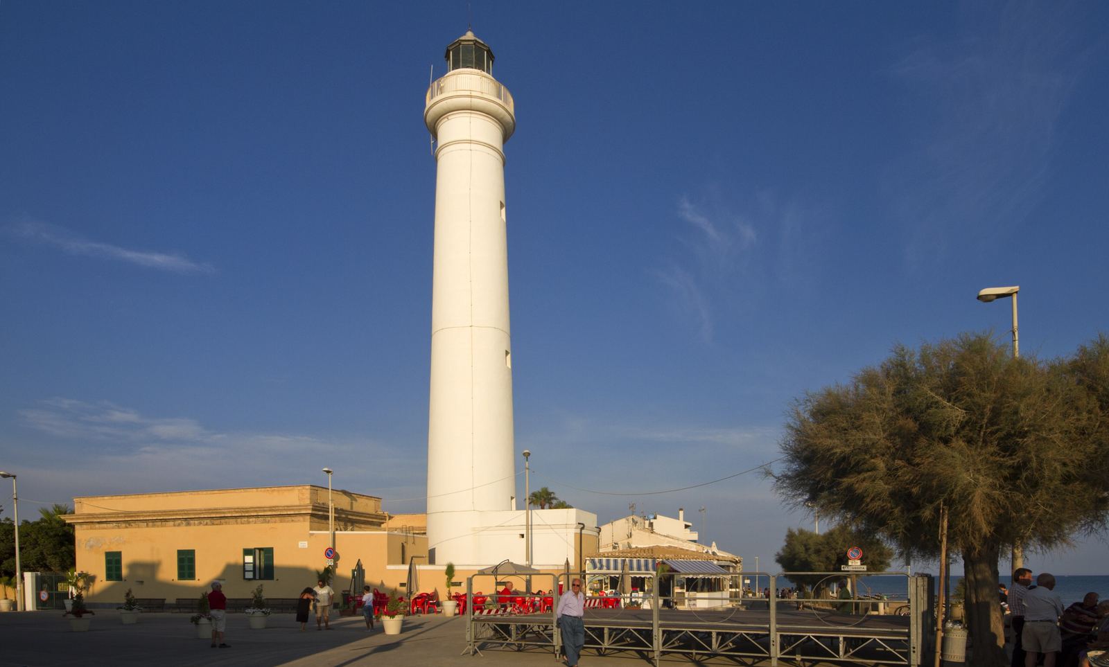 Il faro bianco di Punta Secca con case basse e alberi, sotto un cielo blu.