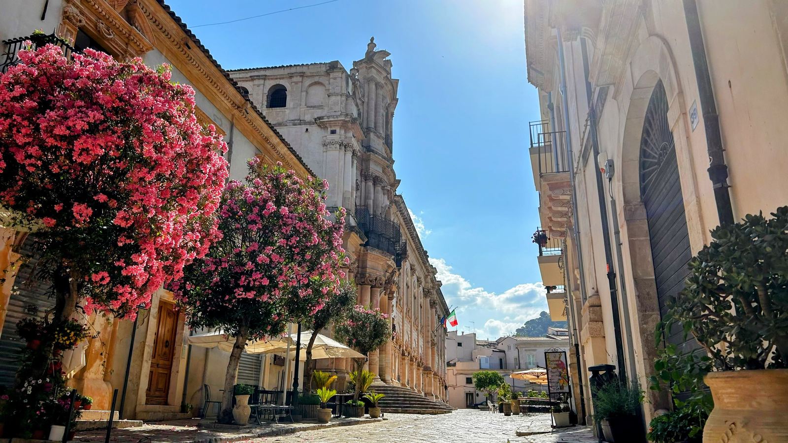 Via Mormino Penna a Scicli con palazzi barocchi, oleandri in fiore e una chiesa in fondo.