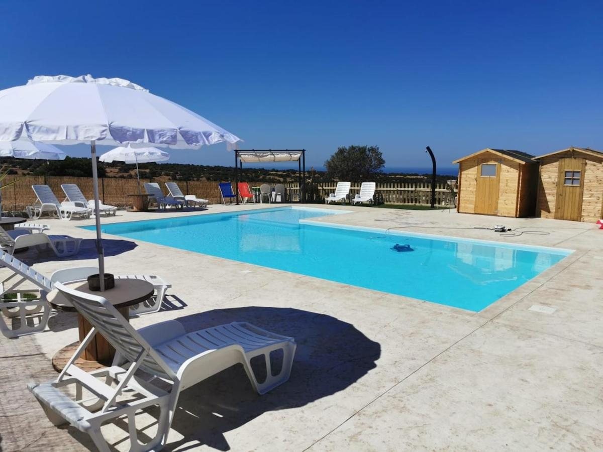 La piscina sotto il cielo azzurro con l'ombrellone bianco, i lettini e le casette in legno